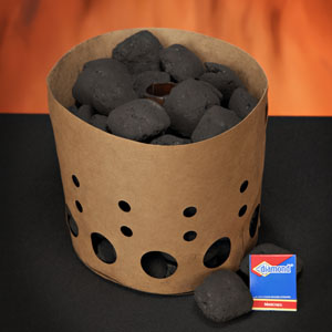 A self-consuming charcoal chimney is loaded with charcoal and sits upon a black surface.