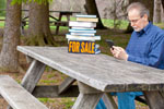 Man reading ebook at picnic table with a for sale sign on the pile of books