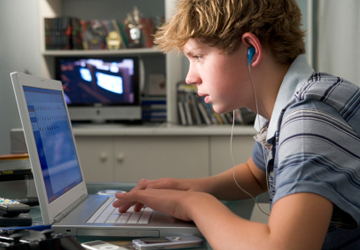 A teenage boy listens to music while using his laptop computer.