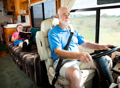 A man drives an RV while his wife sits on a couch behind and reads a book.